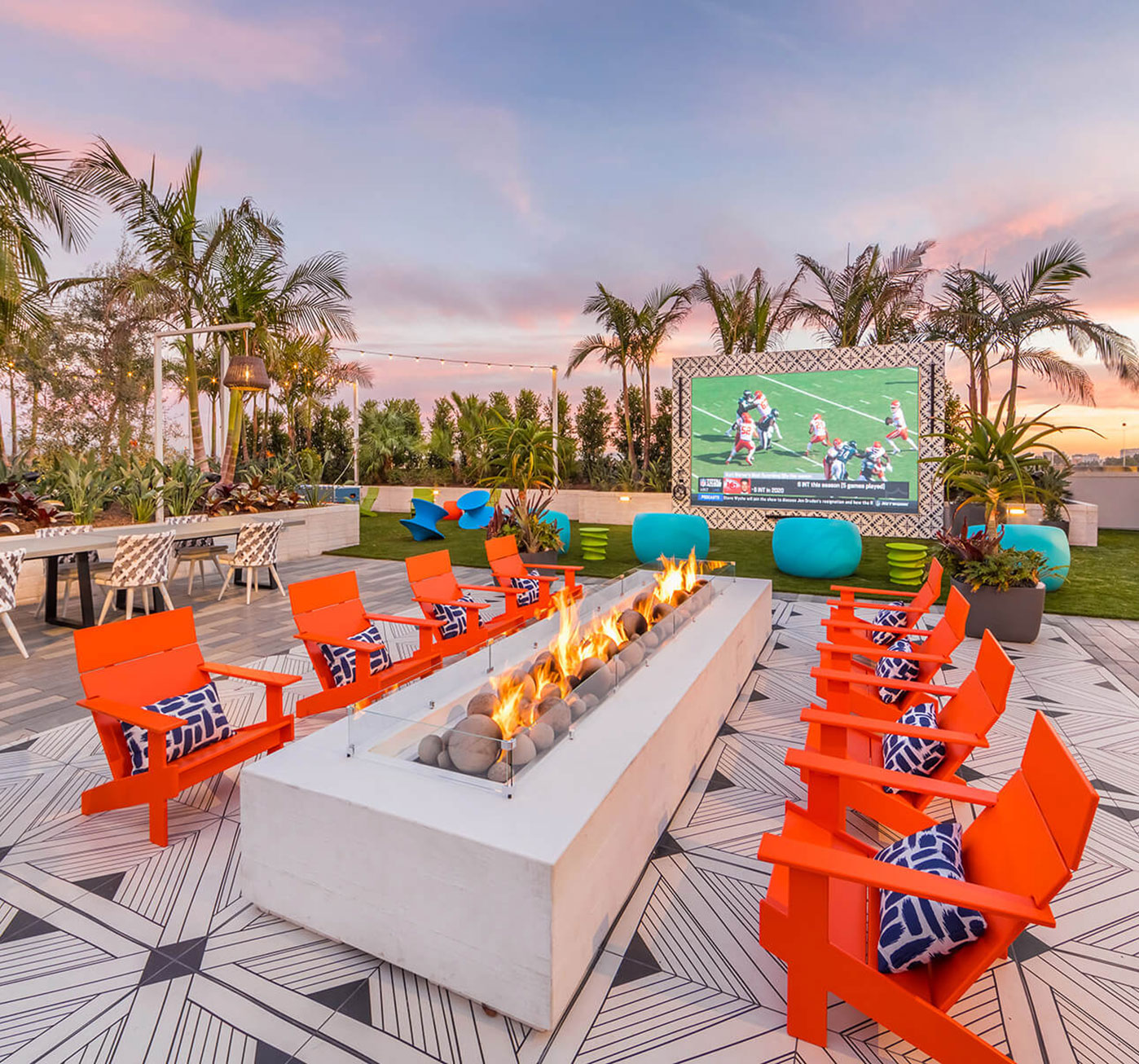 Bright lounge seating around a fire pit sits next to a large video screen on a rooftop terrace lounge surrounded by palm trees.