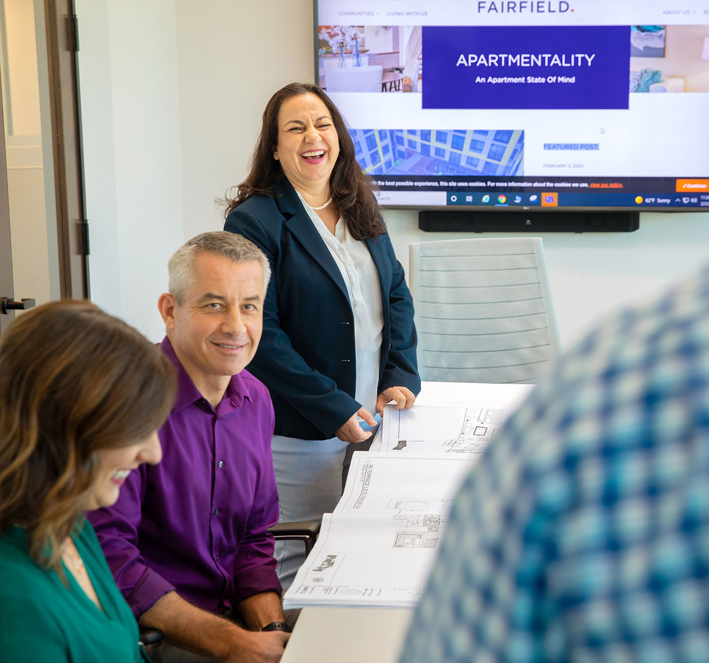 A group of associates laugh during a discussion while looking at construction plan during a meeting in a conference room with a television screen in the background.