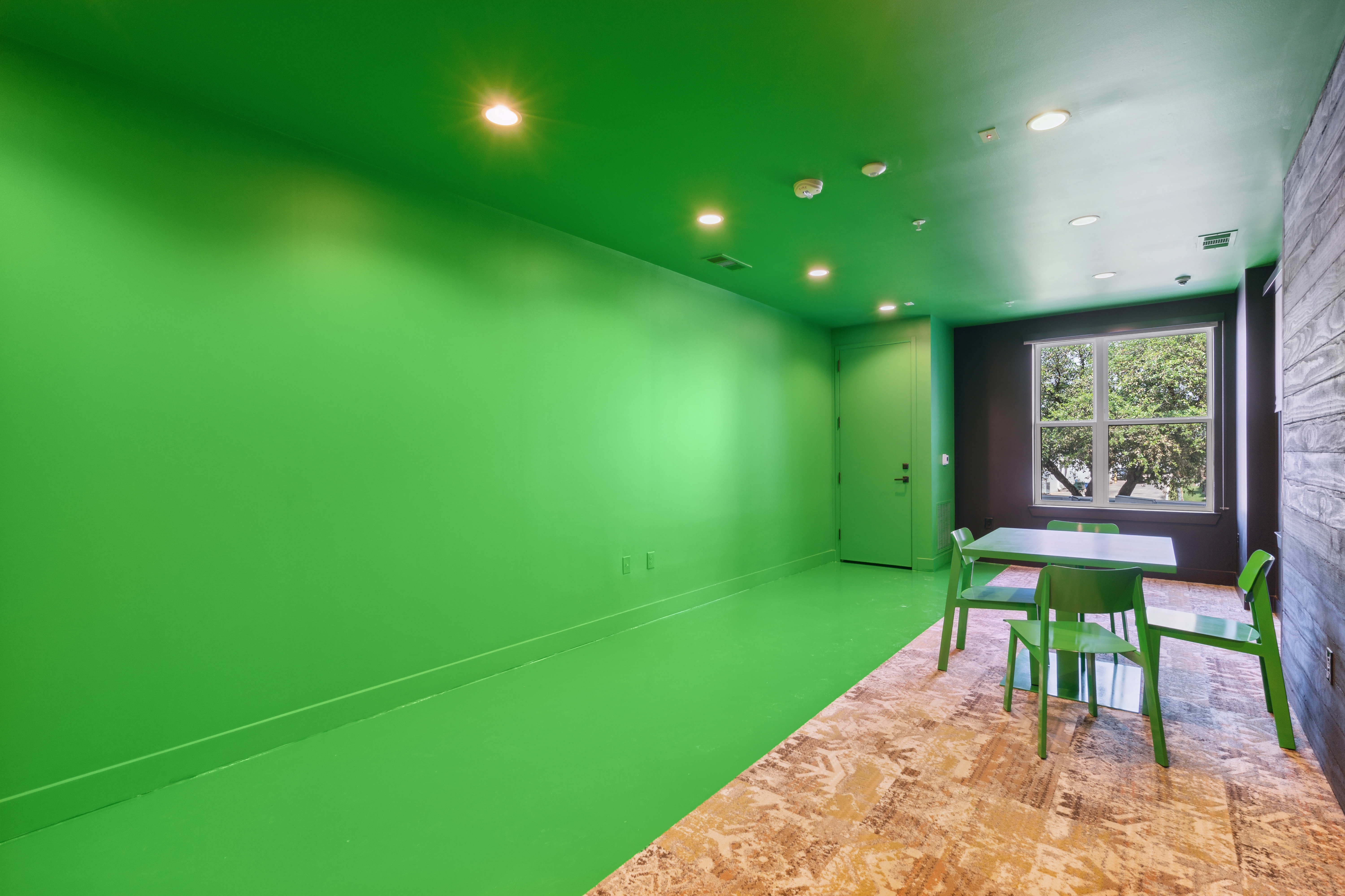 Chroma green covers the floor, ceiling and entire length of the wall until a window looks out at a historic tree. The room includes a chroma green table with four chairs.