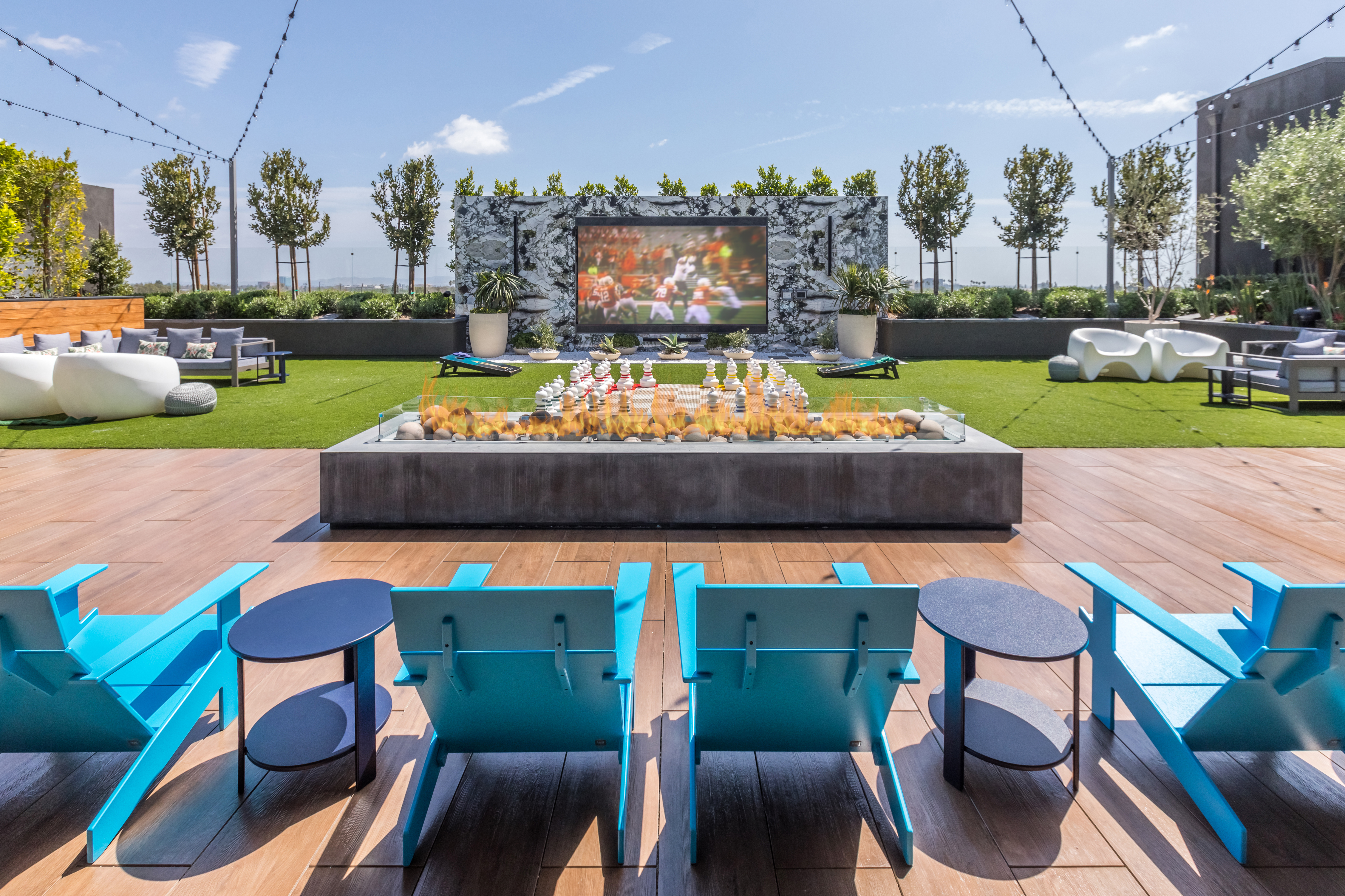 Four adirondack chairs seating before a long concrete fire table overlooking a large movie screen on artificial turf.